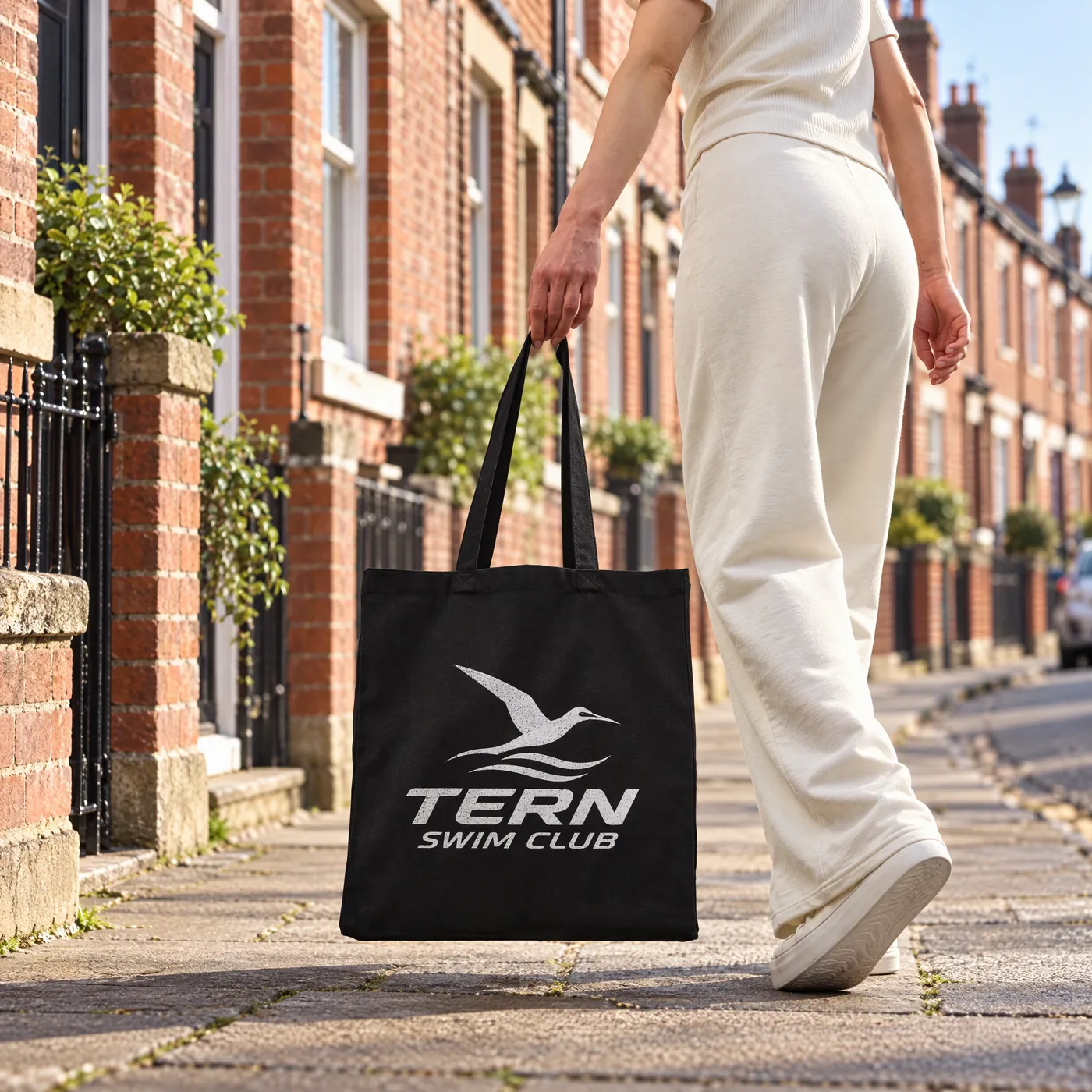 Black recycled canvas tote printed with a swim club logo in white, carried along a terraced street