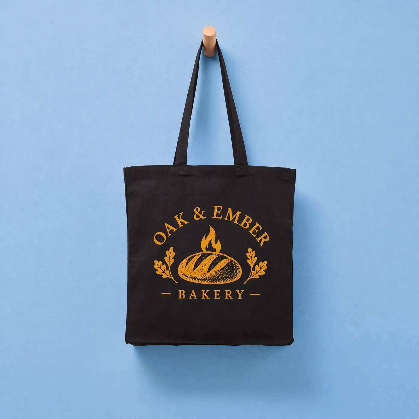 Black recycled canvas tote printed in orange and yellow with an Oak & Ember Bakery logo, hanging on a peg