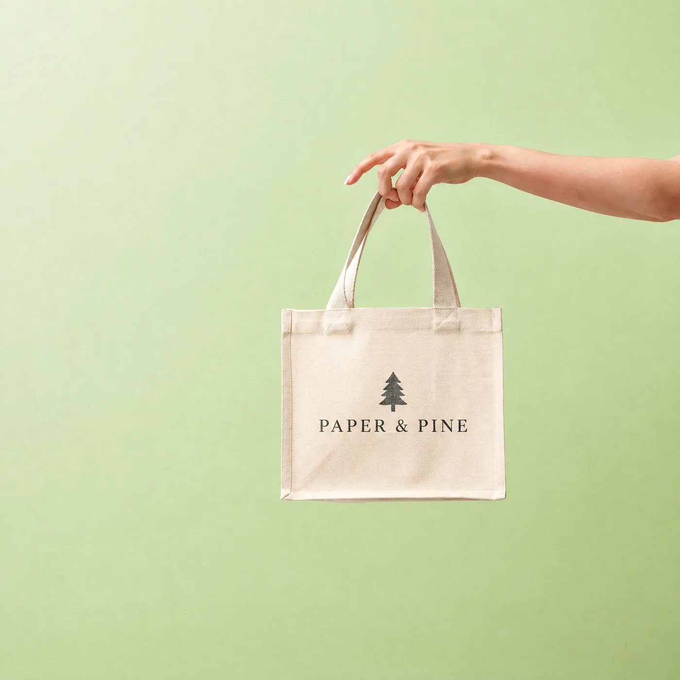 Small natural canvas tote printed with a Paper & Pine logo, held up by its handles