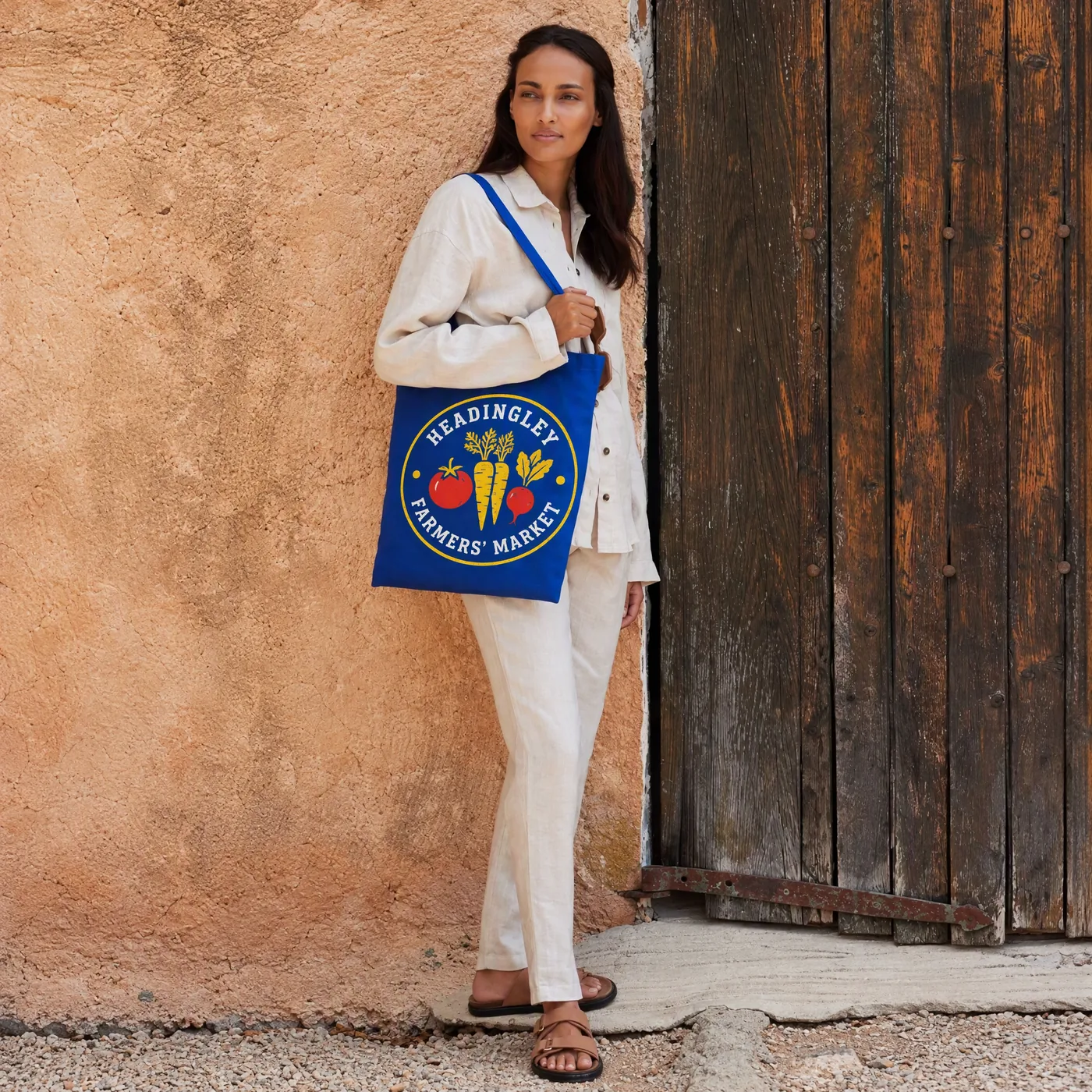 Woman carrying a bright royal blue cotton tote printed with a Headingley Farmers' Market logo, leaning against a sunlit wall