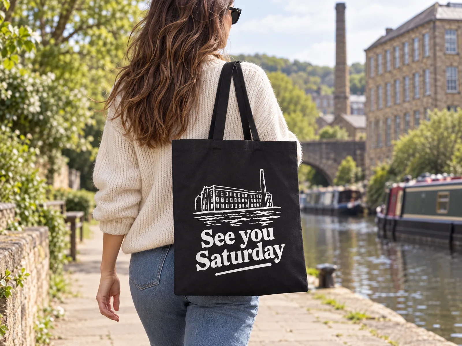 Woman on a canal towpath with a black tote on her shoulder, the back printed with a white See you Saturday slogan