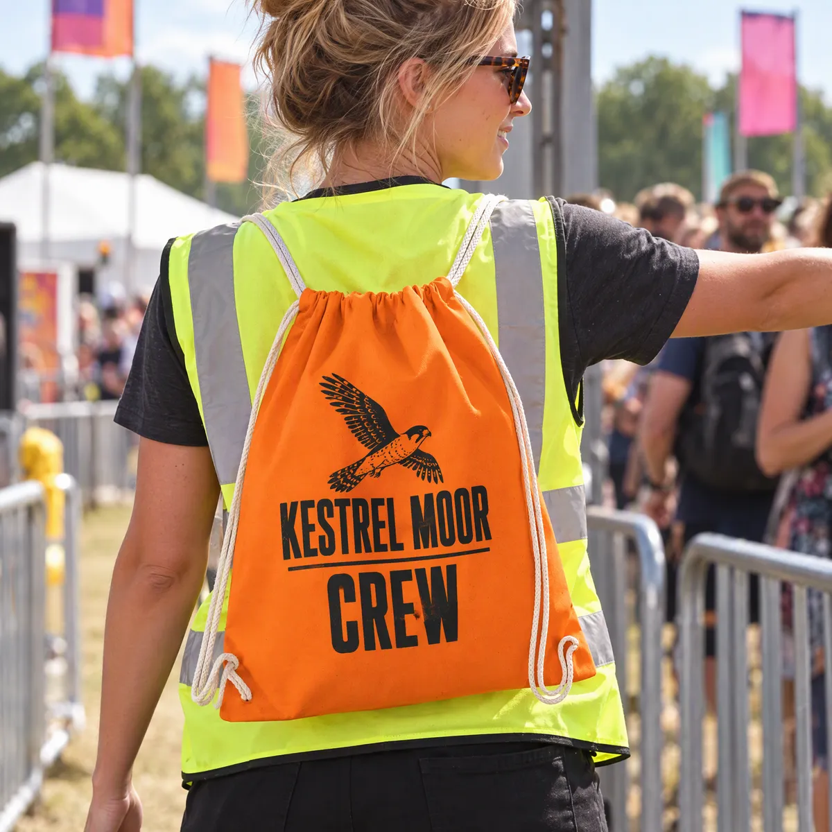 Orange cotton drawstring bag printed with a black Kestrel Moor Crew logo, worn by a festival volunteer