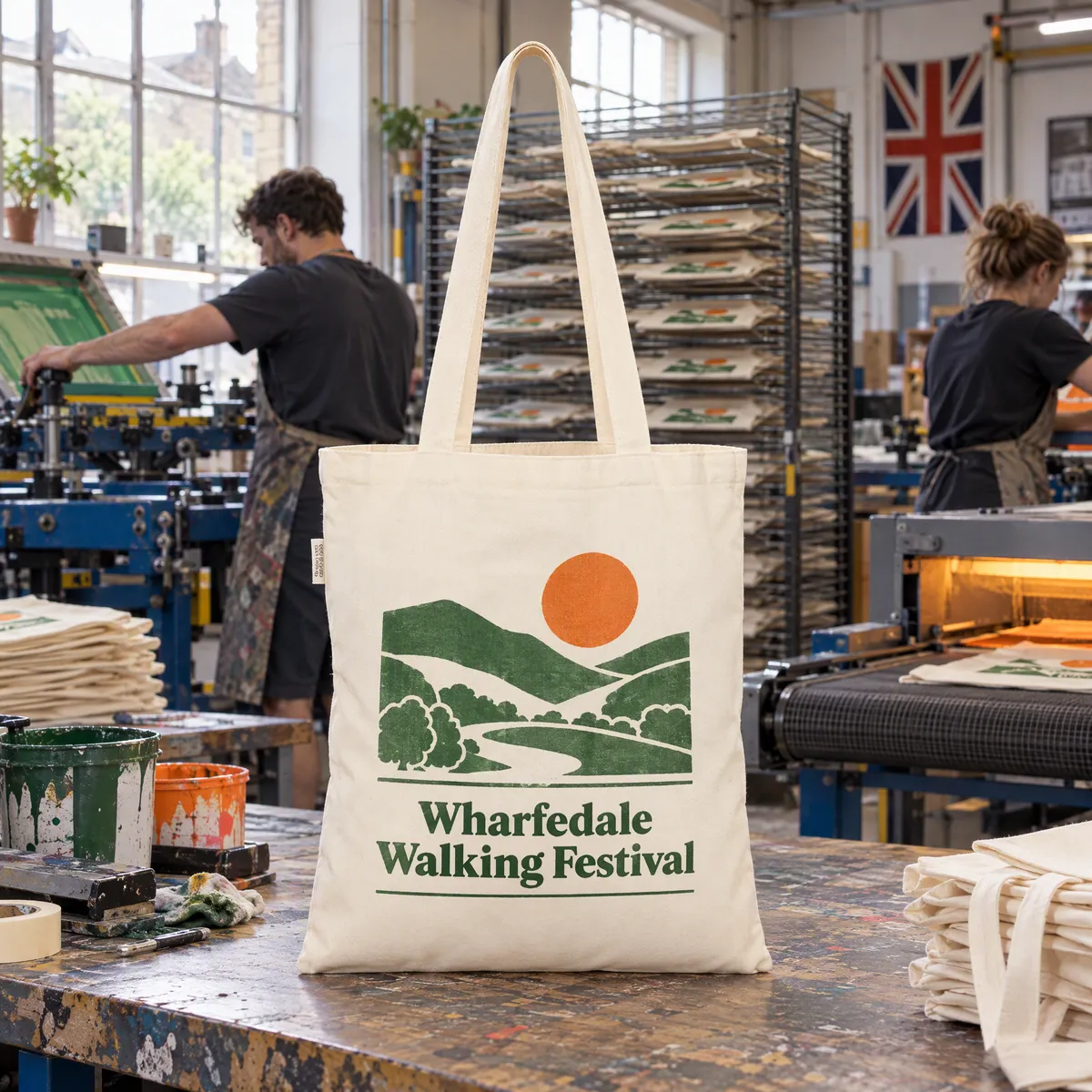 Two printers at screen printing presses with racks of printed tote bags drying behind them