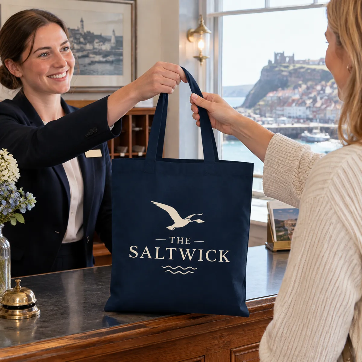 Navy cotton tote printed with a cream Saltwick hotel logo, handed to a guest at a hotel reception desk