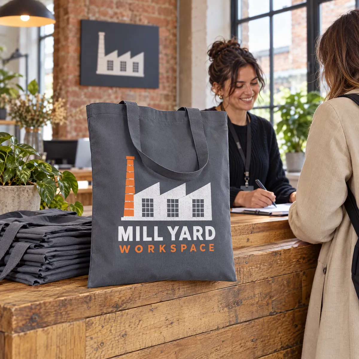Graphite grey cotton totes printed with a Mill Yard Workspace logo, stacked on a coworking welcome desk